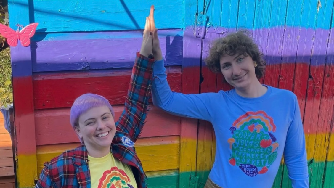 Two people wearing Cooper-Young Community Farmers Garden t-shirts give each other a high five in front of a rainbow wall.